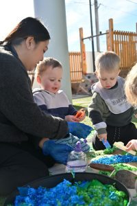 Parent Testimonials - Adult woman helping three young children play with colorful sensory materials at an outdoor table.